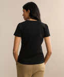 A woman with long dark hair, facing away, wears the black Rib Tee—featuring a fitted body—with tan pants. She stands against a beige background, hands in her pockets.