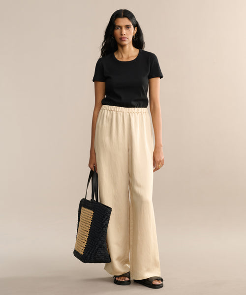 A woman faces forward, wearing a fitted Rib Tee with a U-neck, loose beige pants, black sandals, and holding a large black woven tote bag with a tan panel. The background is plain beige.