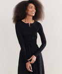 A woman with voluminous curly hair smiles in a fitted Ribbed Finley Cardigan and matching black skirt, channeling a subtle 90s-inspired vibe against a light background.