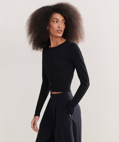A woman with voluminous curly hair models the fitted, long-sleeve black Ribbed Finley Cardigan with high-waisted black pants, standing against a plain background and gazing to the side with one hand in her pocket.