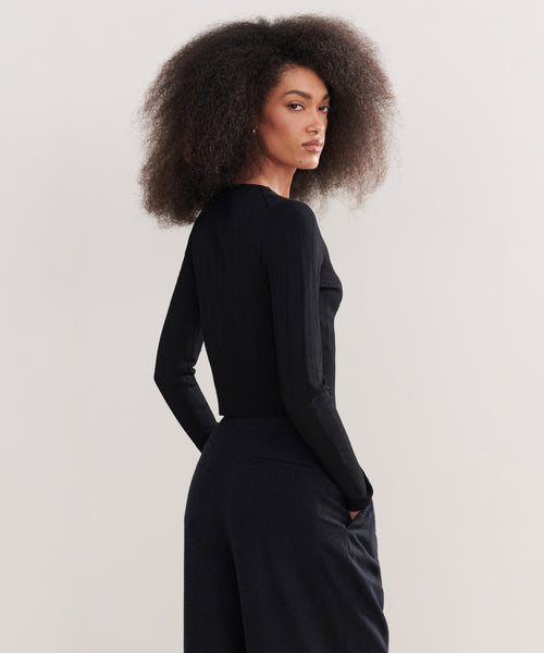 A woman with voluminous curly hair wears the Ribbed Finley Cardigan and black pants, standing with her back slightly turned and hands in her pockets against a plain light background, channeling a 90s-inspired vibe.