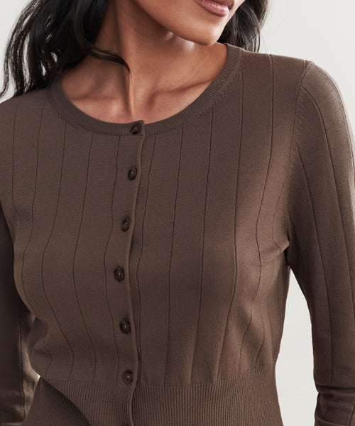 A woman wears the Ribbed Finley Cardigan, a brown, ribbed, long-sleeve button-up in a shrunken 90s-inspired fit. The image is cropped from shoulders to waist, highlighting its textured fabric and distinct buttons.