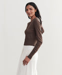 Wearing the Ribbed Finley Cardigan in brown with high-waisted white pants, a person with long dark hair stands sideways against a plain light background, gently smiling and highlighting a subtle 90s-inspired look.