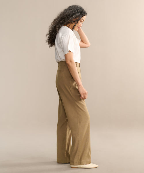 A woman with long curly hair stands in profile against a beige background, wearing a white t-shirt, Riley Trouser—high-waisted lightweight linen trousers—and off-white shoes.
