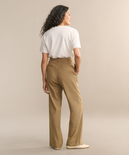 A woman with long curly hair, seen from behind, wears a white t-shirt and the Riley Trouser—a lightweight tan linen wide-leg pant—paired with cream shoes, standing against a plain beige background.