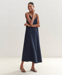 A woman stands smiling in a minimalist setting, wearing the ankle-length Riviera Slip Dress with thin straps and black sandals. Crafted from raw silk, it complements her pulled-back hair and gently crossed arms.