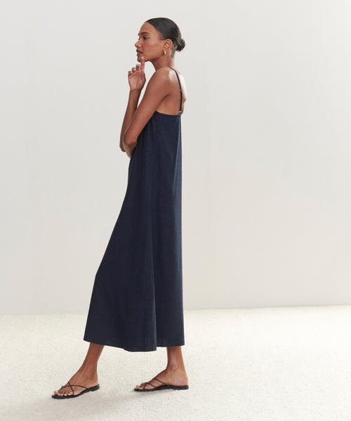 A woman in profile models the Riviera Slip Dress—a loose, sleeveless style with thin straps—paired with black sandals. Her hair is in a bun as she poses with one hand near her face against a light background.