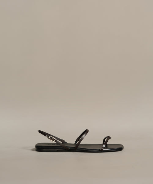 The Romy Sandal is a black minimalist design with thin leather straps and a small gold buckle, handmade in Tuscany and shown in profile against a plain beige background.