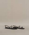 A pair of minimalist Romy Sandal strappy sandals in black, featuring thin straps and handmade in Tuscany, displayed on a neutral beige background.