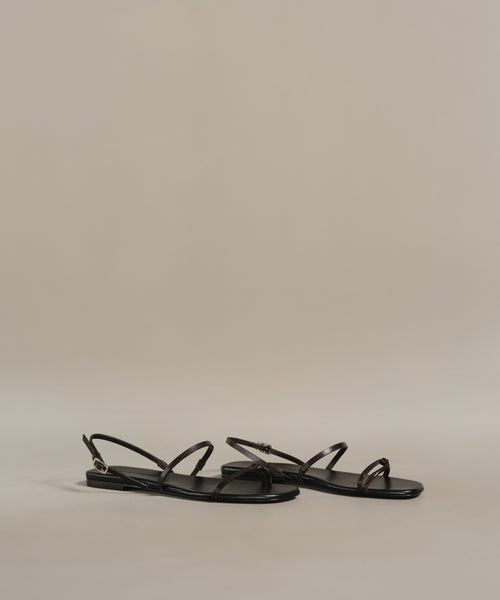 A pair of minimalist Romy Sandal strappy sandals in black, featuring thin straps and handmade in Tuscany, displayed on a neutral beige background.