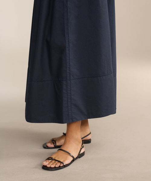 A person in a long, dark navy skirt wears Romy Sandals, handcrafted in Tuscany, standing on a beige floor. Only the lower legs and feet are visible.