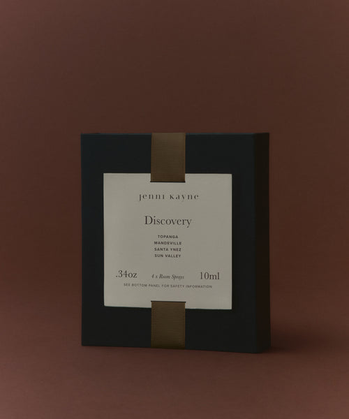 A black Room Spray Sample Scent Set box with a white label featuring "jenni kayne Discovery" and the scent names: Topanga, Mandeville, Santa Ynez, and Sun Valley. Includes four 10ml best-selling room sprays. Brown background.