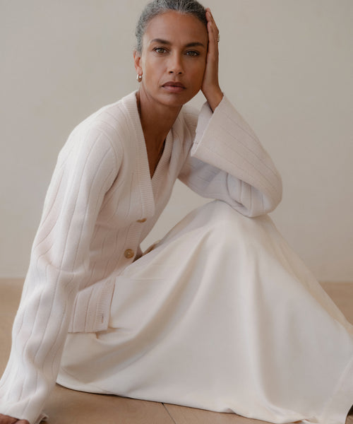 A gray-haired woman sits on the floor in a cream Rory Cardigan with a V-neck, styled with a long white skirt. She rests her head on her hand and looks calmly at the camera, set against a neutral background.