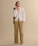 Against a beige backdrop, a person wears the Rory Cardigan—a light beige V-neck knit with large buttons—styled with tan wide-leg trousers, white shoes, and a brown coat draped over their shoulder, looking thoughtfully to the side.