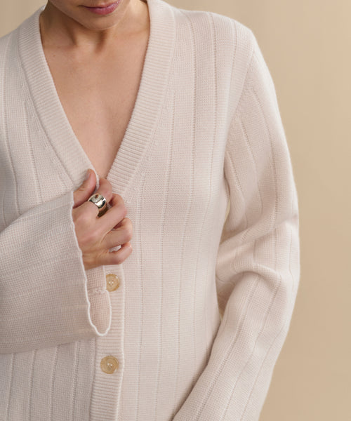 A person wears the Rory Cardigan, a cream ribbed V-neck knit with large buttons, slightly open at the chest. They hold it closed with one hand, showing a silver ring against a neutral beige background.