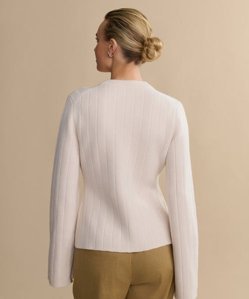A woman with blonde hair in a neat bun is shown from behind, wearing the Rory Cardigan— a fitted, long-sleeve V-neck light pink ribbed sweater—paired with tan pants against a plain beige background.