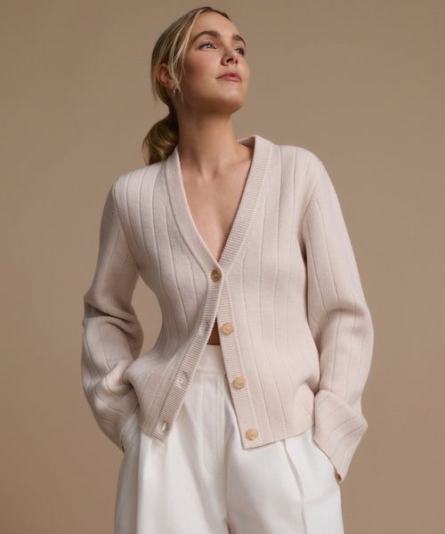 A woman in the Rory Cardigan, a light beige V-neck knit with large buttons, pairs it with white high-waisted pants as she stands against a plain beige background, gazing confidently to the side.