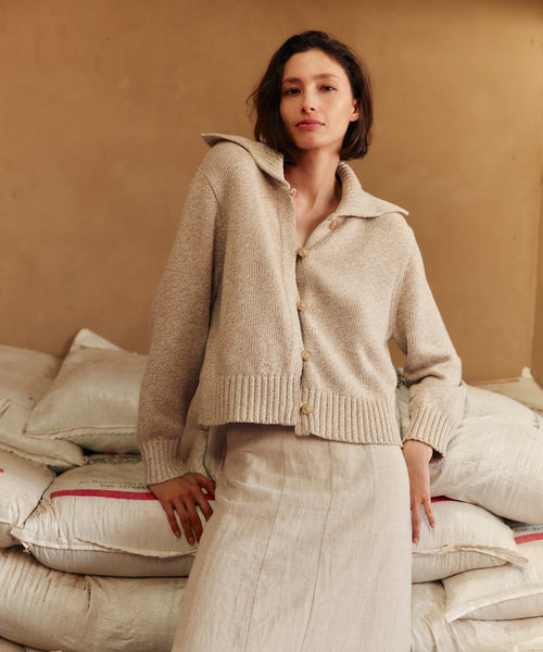 Woman wearing a beige knit cardigan standing among stacked bags against a beige wall.