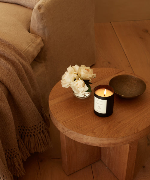 A round wooden side table holds the Santa Ynez Glass Candle, a small glass vase of white roses, and a brown bowl next to a beige sofa with a brown fringed throw blanket on a wooden floor.