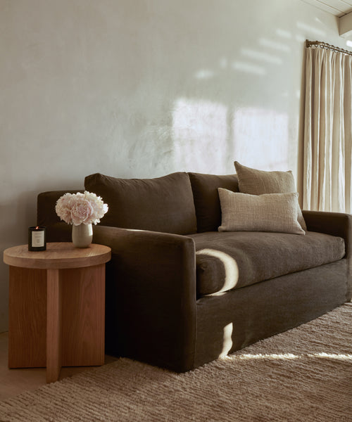A cozy living room corner with a dark brown sofa, beige pillows, and a wooden side table featuring a Santa Ynez Glass Candle and white flowers. Sunlight streams in, casting shadows on the light-colored wall and floor.
