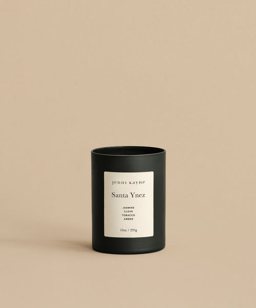 A black Santa Ynez Glass Candle with a white label reading “jenni kayne Santa Ynez, JASMINE, TOBACCO, AMBER, 10oz / 255g” sits on a beige background—a rustic scented candle hand-poured in Los Angeles.