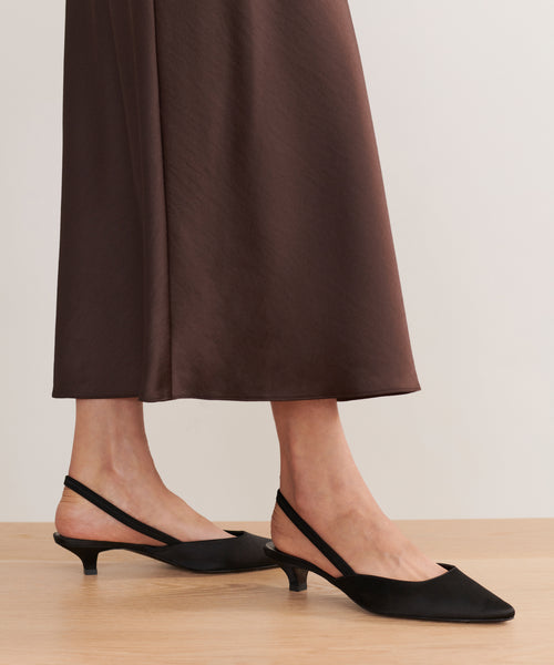 A person stands on a wooden floor against a neutral background, wearing a brown ankle-length skirt and the Satin Kitten Heel—elegant black satin heels made in Italy.