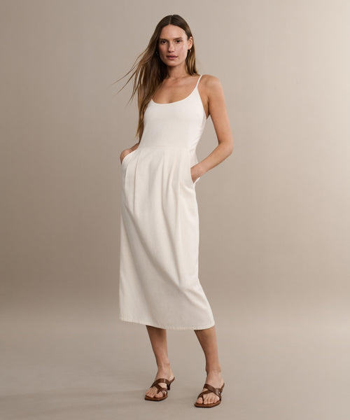 A woman stands against a beige background wearing a sleeveless white midi dress with pockets and the Scarlett Heeled Sandal. Her long, straight hair and relaxed pose add to her confident, summery style.