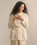 A woman with long curly hair smiles while looking down, wearing the Scout Jacket—a lightweight wool style with a stand collar, large front pockets, and a tie waist—paired with matching pants against a plain beige background.