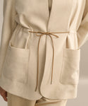 A close-up of a person wearing the Scout Jacket—a beige, lightweight wool piece with large front pockets, a stand collar, and a thin tan drawstring bow at the waist—an effortless wardrobe essential.