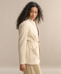 A woman with long curly hair wears the cream-colored Scout Jacket—lightweight wool with a stand collar and drawstring waist—paired with a matching skirt, standing against a plain beige background.