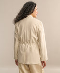 A person with long, curly hair is shown from the back, wearing the beige Scout Jacket with a stand collar and cinched waist, paired with matching pants against a plain beige background.