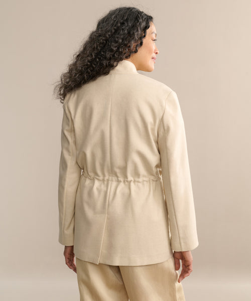 A person with long, curly hair is shown from the back, wearing the beige Scout Jacket with a stand collar and cinched waist, paired with matching pants against a plain beige background.