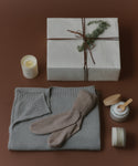 A cozy flat lay displays a folded gray sweater, beige socks, a lit candle, a cream jar with spatula, and the Seasonal Essentials Gift Box tied with string and greenery, all arranged on a warm brown background.