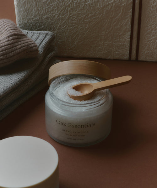 A glass jar of Oak Essentials Dream Bath Soak with a wooden lid and spoon, arranged beside knitwear and textured boxes—a soothing self-care treat included in the Seasonal Essentials Gift Box.