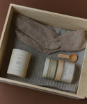 The Seasonal Essentials Gift Box features a wooden box with beige wool socks, a gray knit sweater, a white cedar-scented candle, two Oak Essentials skincare jars, and a small wooden spoon—all elegantly arranged.