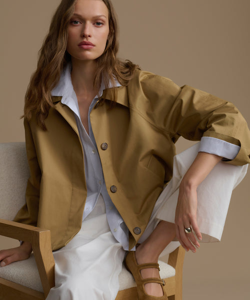 Woman wearing a mustard yellow jacket over a white shirt, sitting on a chair against a beige background.