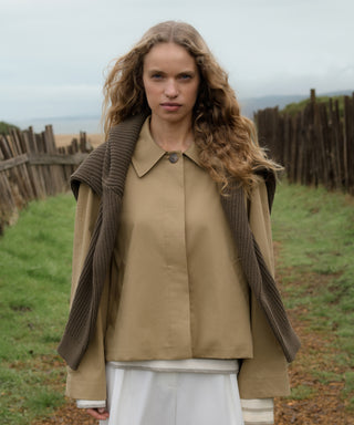 Woman wearing a beige coat with a brown sweater over it, standing in a field.