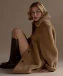 A woman with short blonde hair sits on the floor wearing the Shearling Aurora Coat and knee-high black boots, looking at the camera against a neutral background. The coat adds texture and warmth to her modern style.