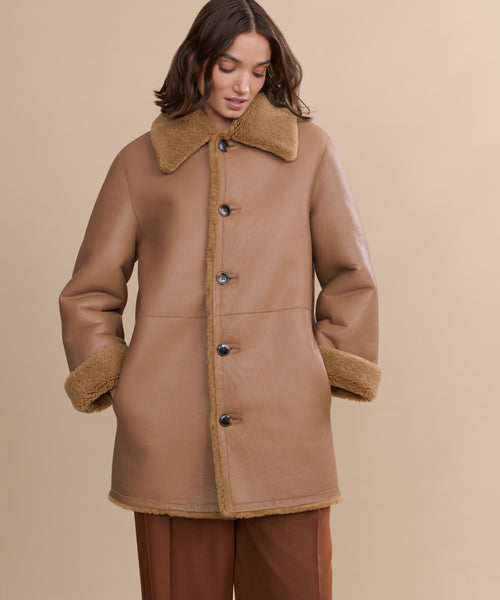 A woman models the Shearling Aurora Coat, featuring a wide collar and large buttons, with her hands in her pockets. Set against a beige backdrop, she highlights the cozy and timeless charm of this reversible tan coat.