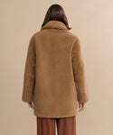 A person with long brown hair is seen from behind, wearing the Shearling Aurora Coat in light brown and brown pants, standing against a neutral beige background.