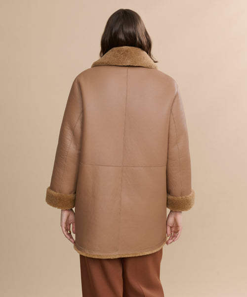A person with medium-length brown hair, seen from behind, wears the Shearling Aurora Coat with turned-up cuffs and matching brown pants against a beige background.