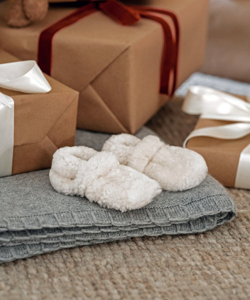 Shearling Baby Booties in ivory rest on a folded gray blanket, surrounded by brown gift boxes with white and red ribbons, all placed on a textured beige carpet.