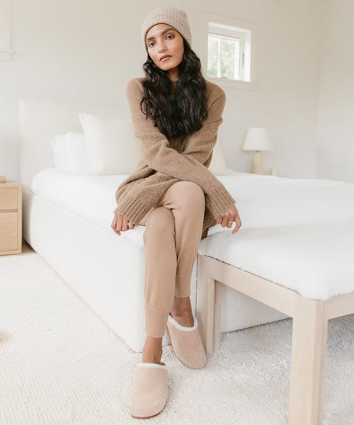 A woman in a beige knit hat, brown sweater, tan pants, and Shearling-Lined Moc Clog sits on the edge of a white bed in a softly lit, minimalist bedroom.