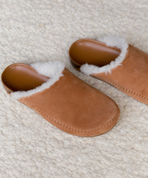 A pair of Shearling-Lined Moc Clogs in tan suede with white shearling lining, handmade in Italy, rests on a textured cream-colored carpet.