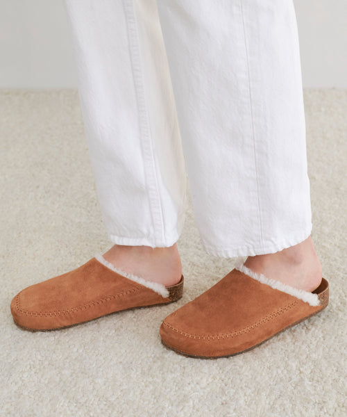 A person stands on a light carpeted floor, wearing white pants and the Shearling-Lined Moc Clog in tan suede with a cork sole.