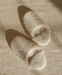 A pair of Shearling Moroccan Slippers in cream rests on a textured beige rug, with soft sunlight casting gentle shadows nearby.