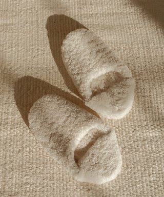 A pair of Shearling Moroccan Slippers in cream rests on a textured beige rug, with soft sunlight casting gentle shadows nearby.