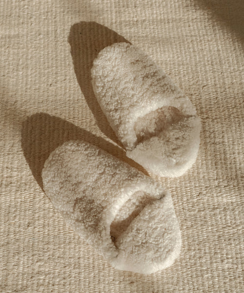 A pair of Shearling Moroccan Slippers in cream rests on a textured beige rug, with soft sunlight casting gentle shadows nearby.