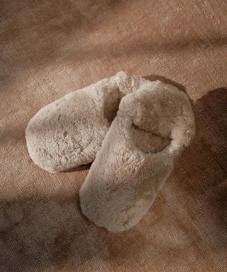 A pair of Shearling Moroccan Slippers in beige with fluffy uppers and soft suede outsoles rest on a textured brown surface, gentle shadows falling across them.