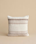 The Sheridan Pillow, a square cushion featuring horizontal beige and light brown stripes on white alpaca blend yarn, is shown upright against a plain beige background.